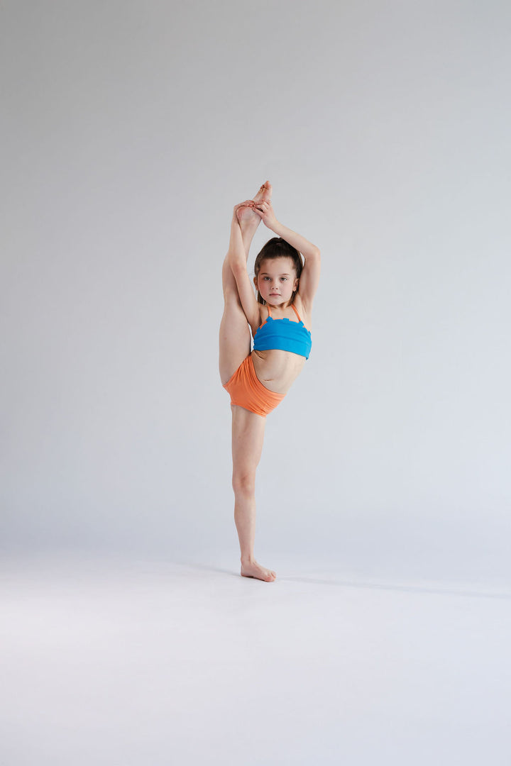 Child performing a split in a blue top and orange shorts on a white background