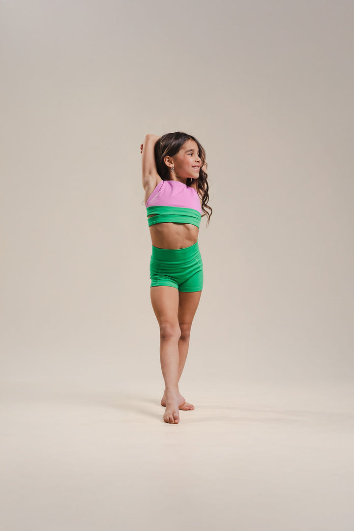 Young girl in a pink and green top with green shorts standing on a beige background