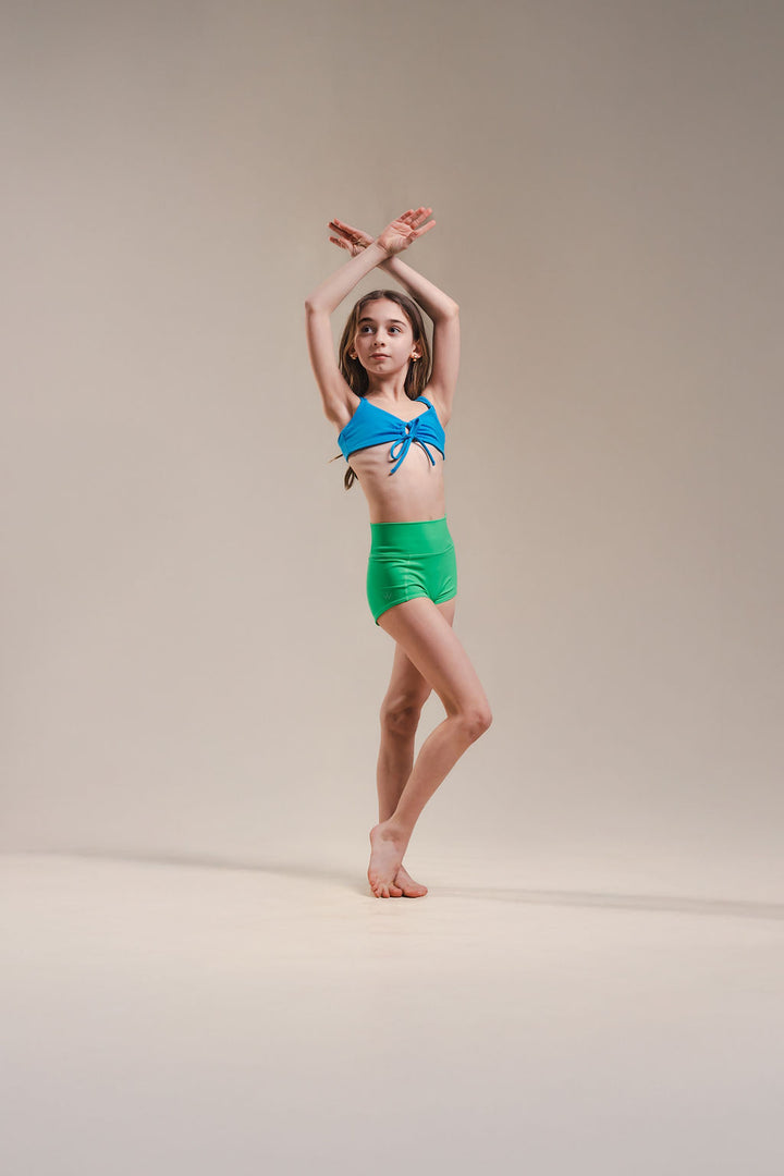 Young girl in a blue top and green shorts posing against a beige background