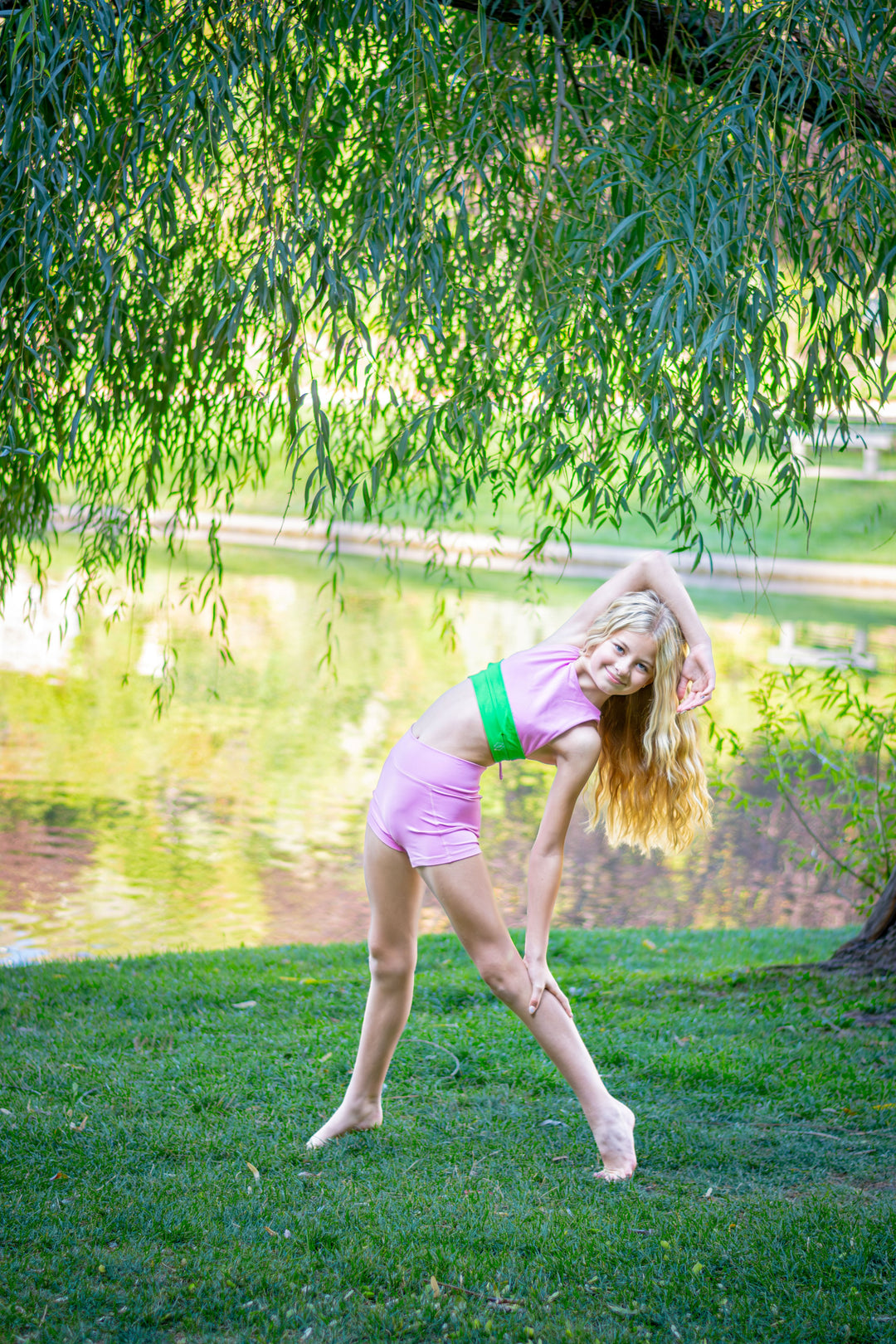 Young girl in a pink outfit standing on grass near a pond with trees in the background